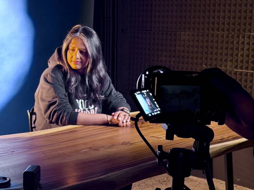 Nida at a studio table during a filmed interview, camera in the foreground